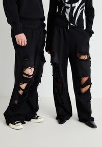 Sorte sweatpants med store slidte huller, der har en løs pasform og frynsede kanter. Båret med kontrastfødder: sorte sneakers og elegante hæle.