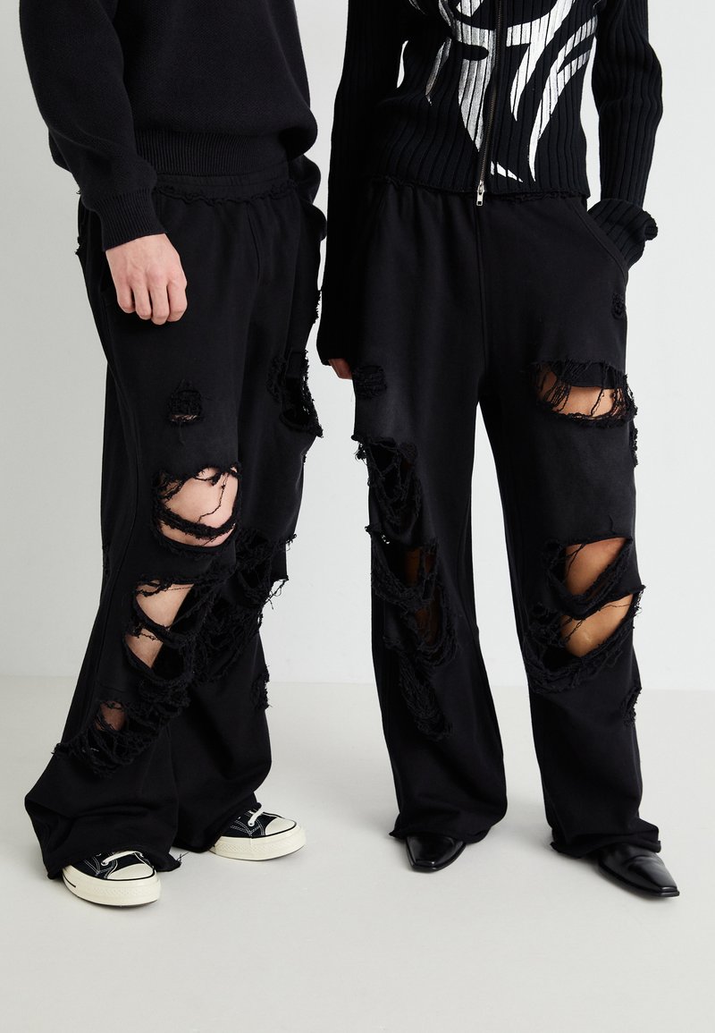 Sorte sweatpants med store slidte huller, der har en løs pasform og frynsede kanter. Båret med kontrastfødder: sorte sneakers og elegante hæle.