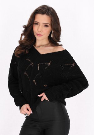 Zwarte gebreide trui met een oversized, off-the-shoulder ontwerp, voorzien van een textuurpatroon en ribgebreide manchetten. Gecombineerd met zwarte broeken.