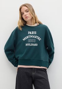 Sudadera de color verde oscuro con el texto blanco "PARIS MONTMARTRE 1993 BOULEVARD", puños acanalados y un dobladillo inferior en capas con rayas. Textura de tela suave.