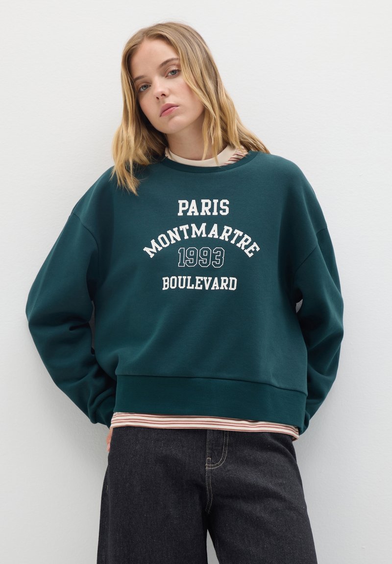 Sudadera de color verde oscuro con el texto blanco "PARIS MONTMARTRE 1993 BOULEVARD", puños acanalados y un dobladillo inferior en capas con rayas. Textura de tela suave.