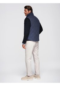 Gilet trapuntato blu navy sopra un maglione nero a trecce, abbinato a pantaloni beige chiaro e sneakers beige. Il modello è mostrato di spalle.
