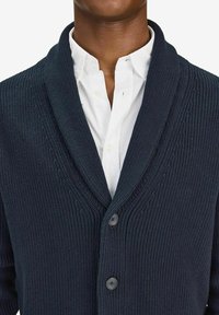 Marinblå ribbade stickade cardigan med sjalkrage, prydd med två knappar, över en vit skjorta med krage. Textur detaljer synliga.