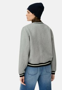 Graue Bomberjacke mit schwarz-weißen gestreiften Details, strukturierter Stoff und taillierte Passform. Kombiniert mit blauen Jeans und einer marineblauen Mütze.
