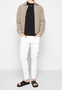 Veste zippée couleur bronze sur un t-shirt noir, pantalon blanc ajusté, et sandales noires. Caractéristiques comprenant un col et des éléments de design minimalistes.