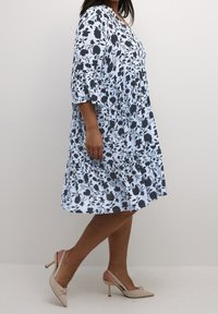 Frau in einem knielangen hellblauen Kleid mit schwarzem Blumenmuster und beigen Slingback-Absätzen, die vor einem einfarbigen Hintergrund geht.