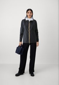 Giacca leggera nera con cerniera e cappuccio giallo a contrasto. Il modello indossa pantaloni neri e tiene in mano una borsa navale con texture.