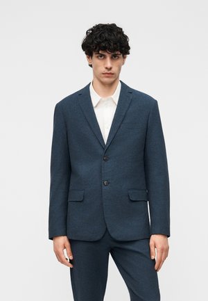 Νεαρός άνδρας με σγουρά μαλλιά, φορώντας εφαρμοστό σκούρο μπλε σακάκι κοστουμιού με δύο κουμπιά και ταιριαστό παντελόνι πάνω από λευκό πουκάμισο με γιακά.