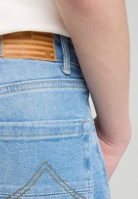 Hellblaue Jeans aus Denim mit einem Lederpatch, das ein Flaggenmuster zeigt. Enthält gelbe Nähte in der Nähe der hinteren Taschen.