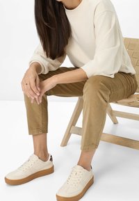 Sweat-shirt crème, pantalon en velours côtelé marron et baskets beiges avec une semelle en gomme et des accents imprimés léopard. La personne est assise sur une chaise en bois.