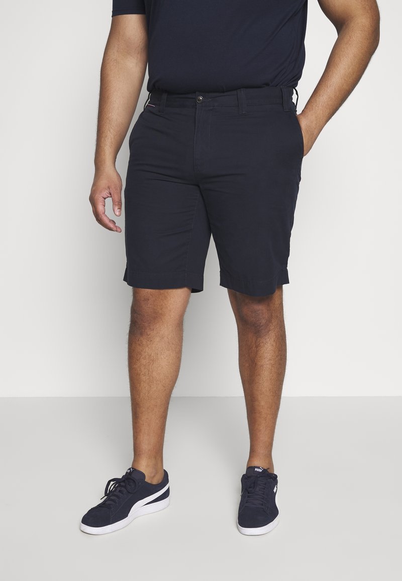 Homme portant un short bleu marine, des baskets bleu marine à semelles blanches, debout avec une main dans la poche devant un fond uni.