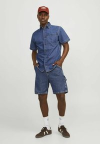 Camisa de denim de manga corta, con un bolsillo en el pecho, combinada con pantalones cortos de denim hasta la rodilla. Zapatillas marrones y una gorra roja completan el conjunto.