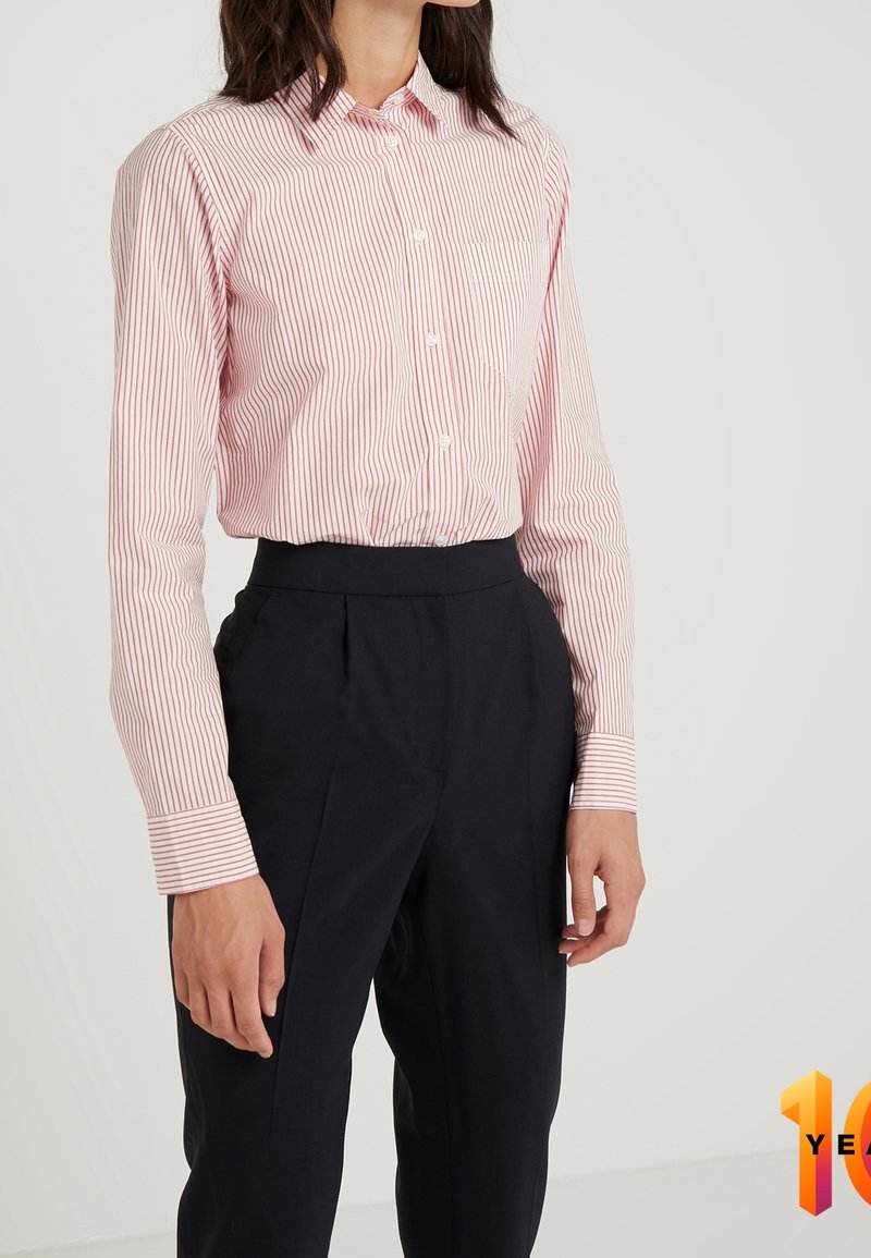 Vrouw in een roze-wit gestreepte button-up shirt, gestoken in hoge zwarte op maat gemaakte broeken tegen een eenvoudige achtergrond.
