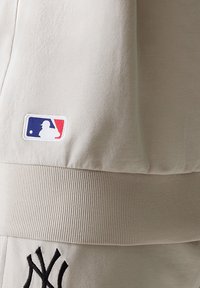 Abbigliamento MLB beige chiaro caratterizzato da un tessuto testurizzato, una vita a coste e un logo ricamato con contorno nero e accenti rossi e blu.