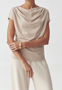 Beige pusero, jossa on cowl-kaulus ja sileä, valuva kangas, lyhyillä hihoilla, yhdistettynä vaaleanbeigeihin leveälahkeisiin housuihin. Kultaista korua korostava yksityiskohta.