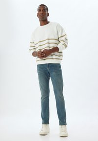 Felpa bianca con strisce orizzontali beige, abbinata a jeans azzurri chiari e scarpe da ginnastica beige. Uomo in piedi su uno sfondo neutro.