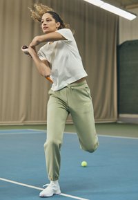 Athletic outfit includes a white short-sleeve shirt and light green joggers, with elastic cuffs. White sneakers are worn, and a tennis racket is held.