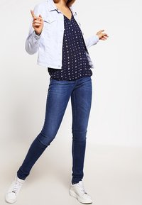 Lyseblå denimjakke over en marineblå bluse med hvidt print, parret med mørke stramme jeans og hvide sneakers. Modellen poserer med den ene hånd hævet.