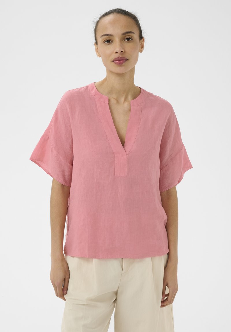 Femme portant un chemisier ample rose à col en V avec des manches courtes et un pantalon beige clair, debout devant un fond blanc.