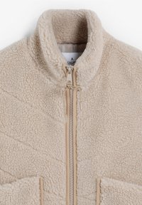 Chaqueta de forro polar beige con un alto cuello, cremallera frontal y dos bolsillos laterales. Presenta un tejido texturizado y detalles de costura en diagonal.