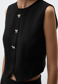Schwarzes ärmelloses Oberteil mit einem cropped Design, glattem Stoff und drei dekorativen silbernen Schleifenakzenten am Frontverschluss.