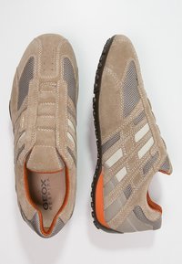 Beige och grå mocka- och nät-sneakers med vita ränder, orange foder och texturerade mörka sulor, visade från ovan och i profil.