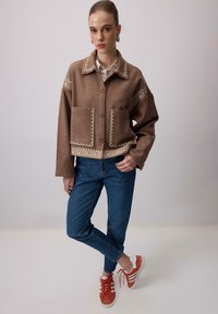 Žena v hnedej bundovej sako s bielou výšivkou, modrých džínoch a oranžových teniskách, stojí s jednou nohou prekríženou cez druhú.