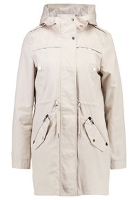 Beige vedenpitävä takki, jossa on huppu, edessä vetoketju ja nepparikiinnitys, kaksi vetoketjullista sivutaskua ja napillinen läppäkoristus.