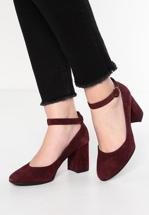 Woman wearing burgundy suede block heels with ankle straps and black cropped jeans with frayed hems on white floor.
