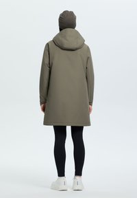 Vihreä vettä hylkivä parka, jossa on huppu, keskikokoinen muotoilu, sileä pinta ja takana tuuletusaukot, yhdistetty mustiin legginsseihin ja valkoisiin lenkkareihin.