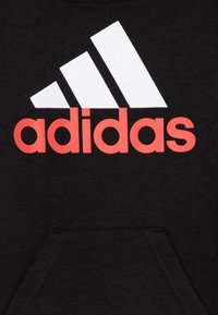 Sudadera negra con un gran logo de tres rayas en blanco y "adidas" en letras rojas brillantes. Cuenta con un bolsillo frontal y una textura suave.