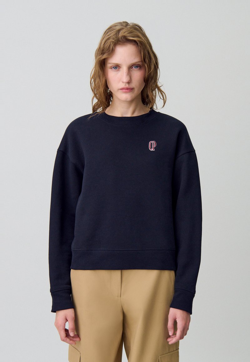 Sudadera negra de algodón con puños y dobladillo de canalé, que presenta un pequeño logo rojo en el pecho. Textura suave y corte relajado.