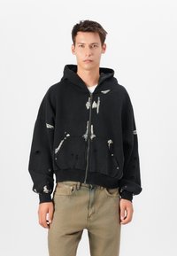 Black zip-up hoodie featuring distressed detailing, including paint splatters and holes. Soft fabric with a relaxed fit. Front pockets included.
