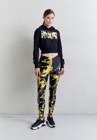 Sudadera corta negra con un logo gráfico, combinada con leggings negros y dorados con un patrón de cadenas y texto. Zapatillas negras completan el look.