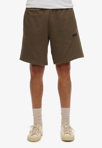 Pantalones cortos de algodón marrón con cinturilla elástica, dobladillo plano y bolsillos laterales. Presenta un logo negro en la pierna izquierda.