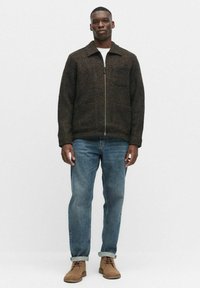 Brązowa kurtka z wełny z zamkiem z przodu, dwoma kieszeniami na piersi, z kołnierzem, zestawiona z niebieskimi dżinsami i brązowymi butami na sznurówki.