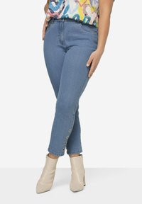 Jeans de mezclilla azul claro con un diseño ajustado, que presentan detalles de botones en el tobillo. Combinados con botas de tobillo beige y una blusa multicolor.
