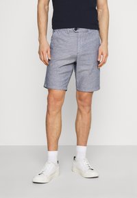 Shorts bleu clair, texturés, en coton. De longueur mi-haute avec une fermeture à bouton à l'avant et des poches latérales. Associés à des baskets blanches et des chaussettes.