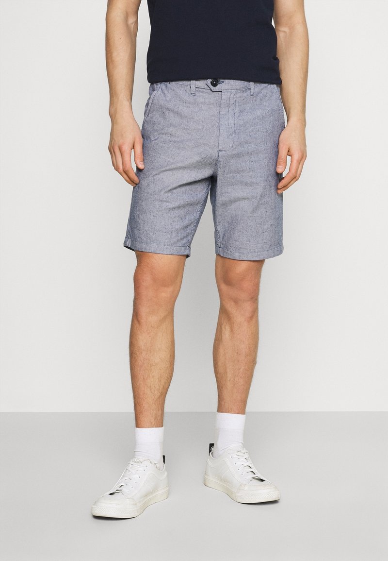 Shorts bleu clair, texturés, en coton. De longueur mi-haute avec une fermeture à bouton à l'avant et des poches latérales. Associés à des baskets blanches et des chaussettes.