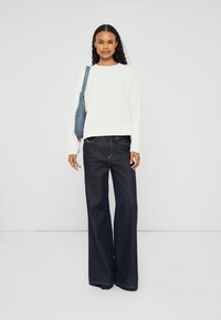 Sweatshirt branco com texto em relevo, combinado com jeans de perna larga azul escuro. Uma bolsa azul está pendurada no ombro. Design simples.
