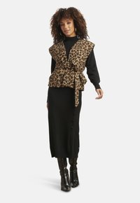 Gilet con stampa leopardata e cintura annodata in vita, indossato sopra un vestito nero a maniche lunghe, abbinato a stivali neri al ginocchio.