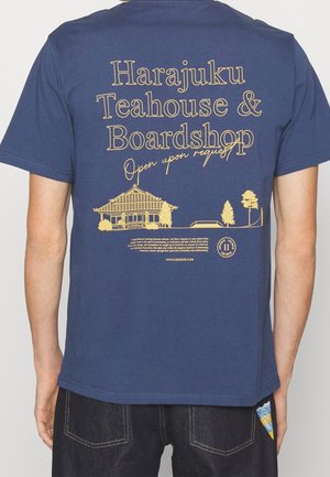 T-shirt en coton bleu marine avec un texte doré "Harajuku Teahouse & Boardshop" et un graphique d'un bâtiment et d'arbres au dos.