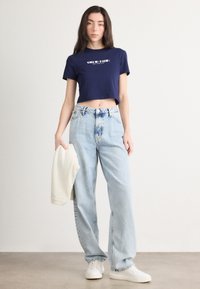 Laivynuosiuotai trumpa marškinė su balta logo, šviesiai mėlyni aukšto liemens džinsai su spalvotu juosmeniu, ir balti sportbačiai, laikantys kreminį megztinį.