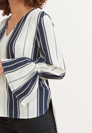 Vrouw draagt een blouse met lange mouwen, brede geplooide klokmouwen en verticale marineblauwe, witte en beige strepen, gecombineerd met een zwarte broek.