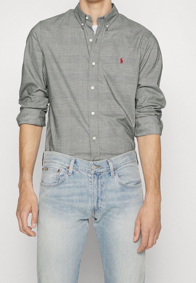 Homme portant un jean bleu clair et une chemise à carreaux grise à boutons avec les manches retroussées et un logo rouge brodé sur la poche poitrine.