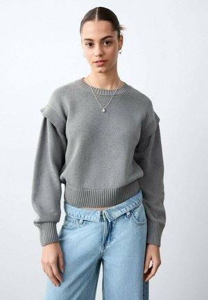 Giovane donna con capelli scuri raccolti in uno chignon, indossa un maglione grigio a maglia e jeans azzurri, in piedi davanti a uno sfondo bianco uniforme.