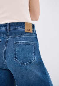 Vaqueros de denim azul con un acabado de lavado medio, una etiqueta de parche en la parte posterior de la cintura y dos bolsillos traseros con detalles de costura visibles.
