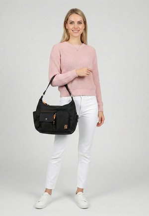 Vrouw in roze trui en witte broek die een zwarte schoudertas met meerdere vakken en oranje ritssluitingen vasthoudt, staand op een effen achtergrond.