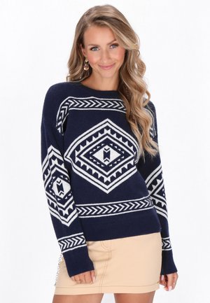 Kvinde iført en mørkeblå sweater med hvide geometriske mønstre og en beige nederdel, smilende med langt bølget blondt hår og øreringe.