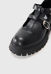 Črne usnjene čevlje s zaobljenim nosom, oblikom T-jermena, podrobnostmi s srebrno sponko in debelim, strukturiranim gumijastim podplatom.